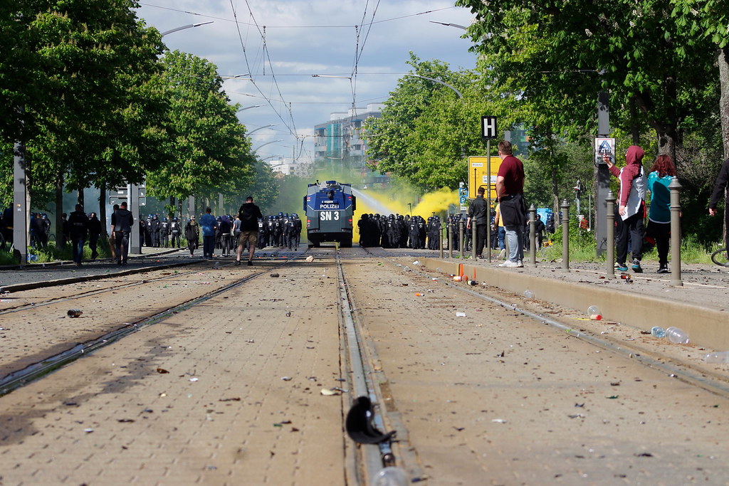 Blick auf die Lennéstraße in Dresden: Im Vordergrund zerbrochene Flaschen, im Hintergrund ein Wasserwerfer im Einsatz, eine Polizeikette und davor gelber Rauch.